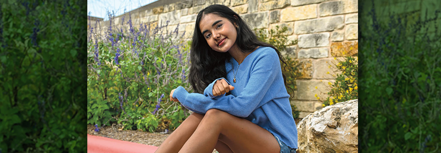 A smiling girl in a blue sweatshirt sits outdoors near a stone wall and purple flowers